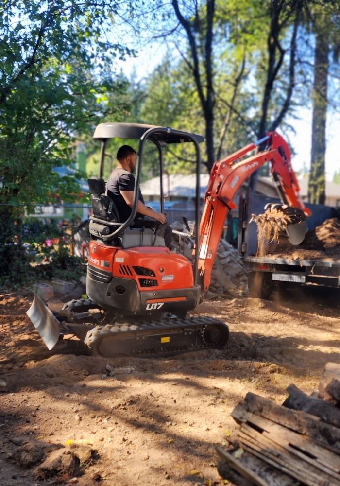 Nate operating a compact excavator to load soil and roots into a dump trailer at a residential job site in Washington